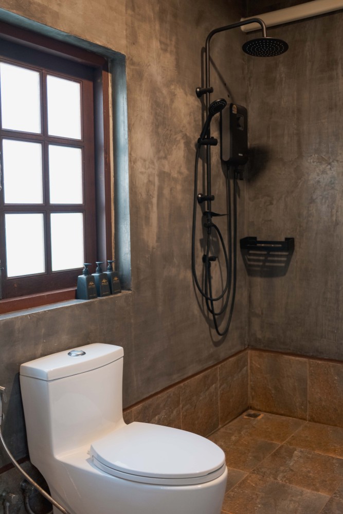 Bathroom at Tree Roots Retreat, Mae Ramphueng, Rayong, with a rain shower, dark polished concrete walls, a teak vanity, and a window onto the plants.