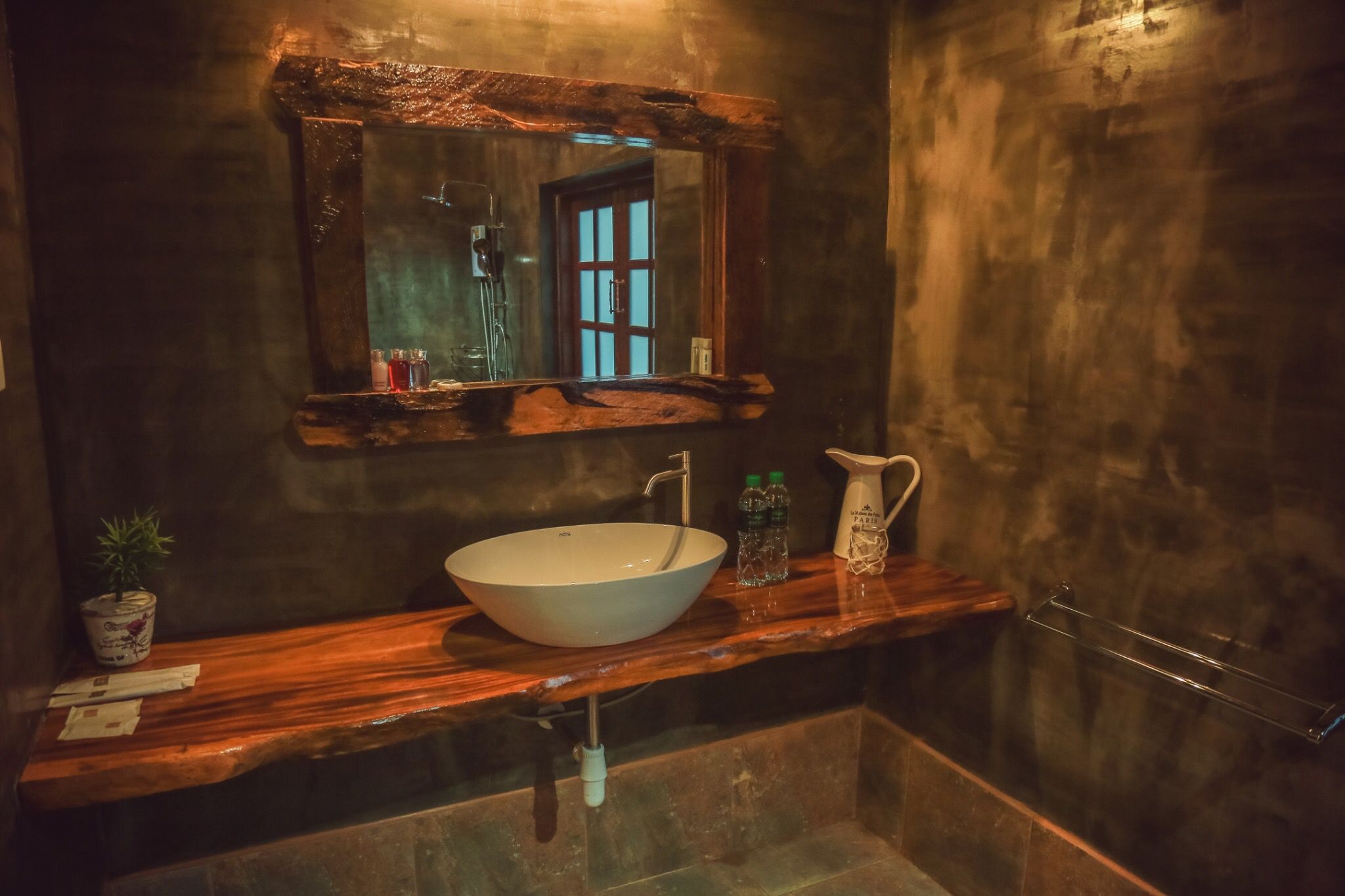 Teak vanity in the bathroom at Tree Roots Retreat, Mae Ramphueng, Rayong, with a white vessel sink, a round mirror, folded towels, and a window onto the plants.