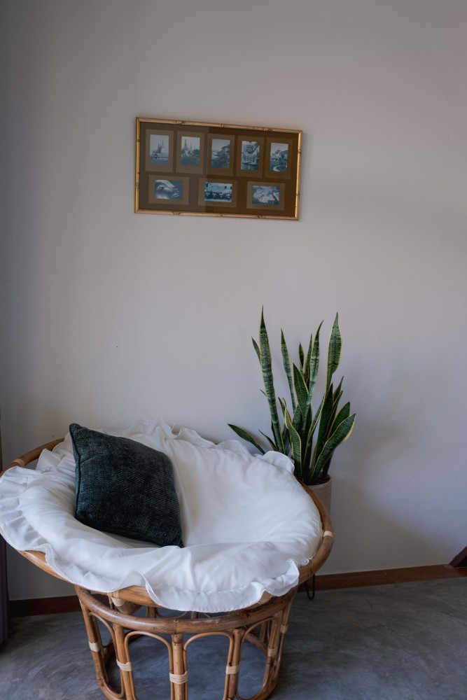 A rattan chair and open laptop in a garden room at Tree Roots Retreat, Mae Ramphueng, Rayong, with books on a side table and the double bed beside a green painting.