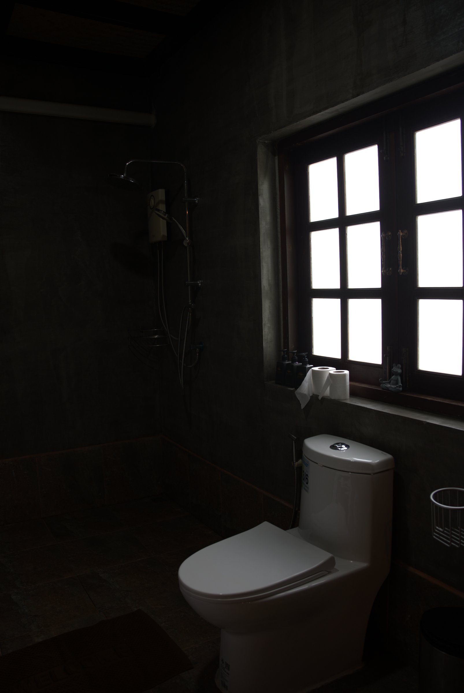 Bathroom in a garden room at Tree Roots Retreat, Rayong, with dark walls, a wood-framed window, and a shower in the background.