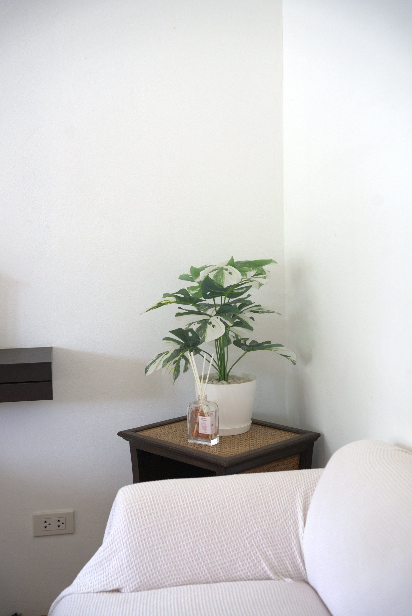 Seating corner in a garden room at Tree Roots Retreat, Rayong, with a white sofa, a rattan-top side table, and a potted monstera.